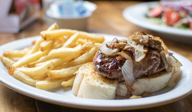 A plate showcasing a toasted baguette with steak on top, topped with onions and mushrooms with a side of fries.  The dining special at The Georgetown Inn and Pub in Canmore, Alberta