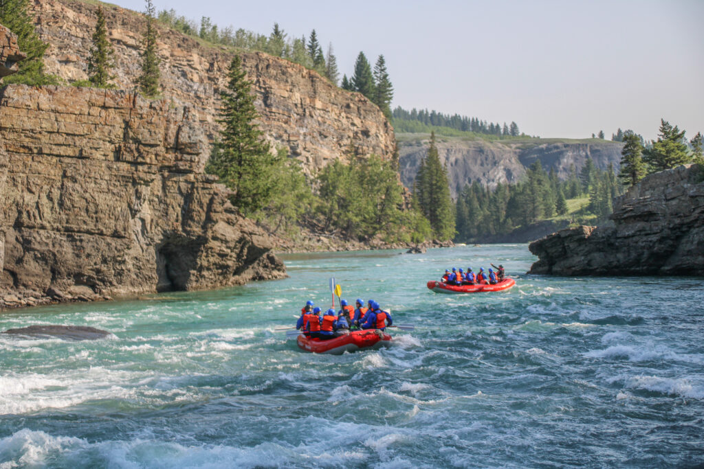 Two red rafts floating down the river surrounded by shale canyon walls, trees and plains