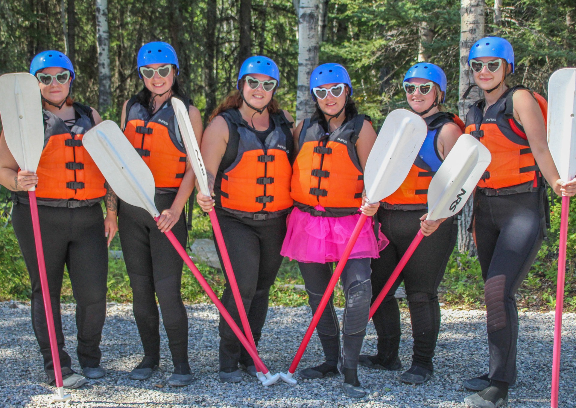 A group of women in matching heart-shaped sunglasses, life jackets, and helmets pose with paddles before a rafting trip, radiating fun and friendship—perfectly capturing the energy of a bachelorette party adventure with Canadian Rockies Rafting.